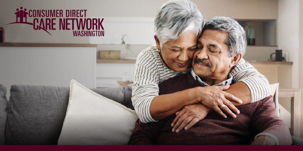 Woman standing behind a couch hugging an elderly man who is sitting on the couch.